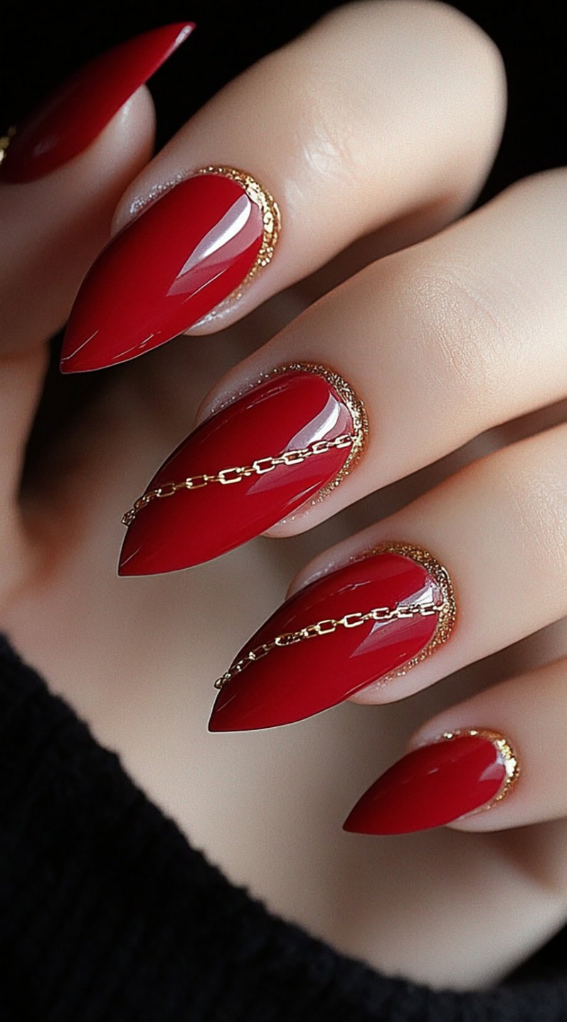 A close-up of stiletto-shaped nails painted in a deep, glossy red with gold glitter accents around the cuticle and delicate gold chains placed across the nails.