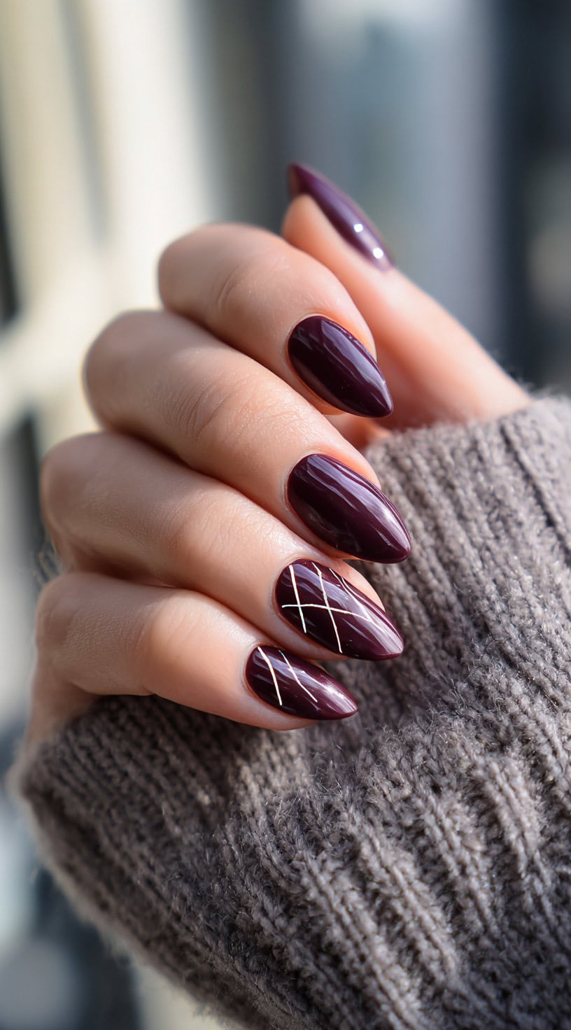 Almond-shaped nails in a glossy wine shade with fine gold criss-cross lines on the ring and middle fingers.
