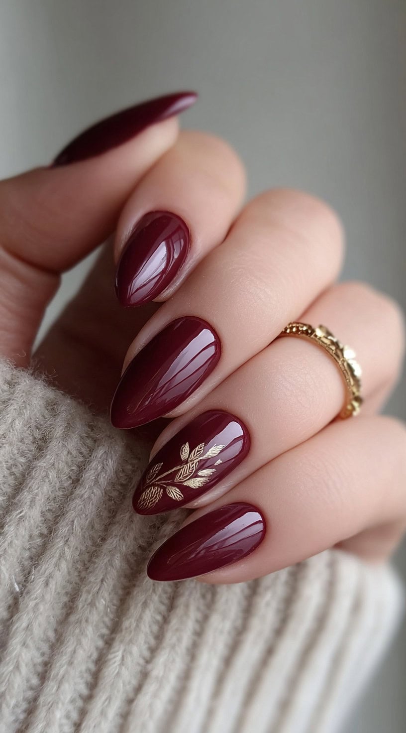 A hand with glossy deep burgundy nails, one of which features a delicate gold botanical decal.