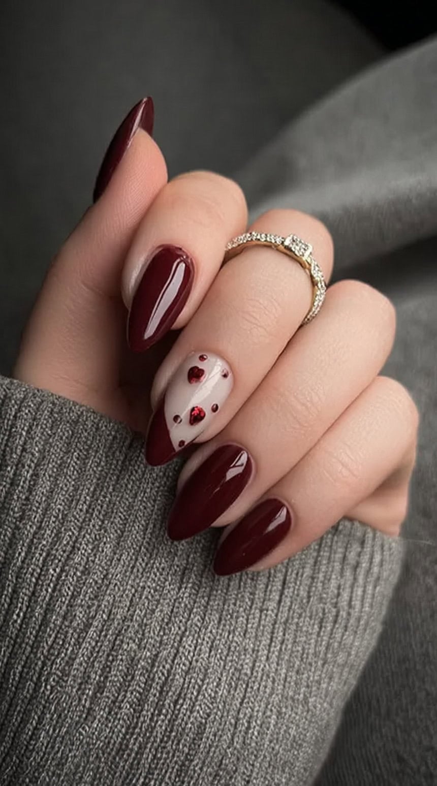 A hand with almond-shaped nails in glossy wine polish, featuring one nude accent nail decorated with red heart-shaped gems and dots.