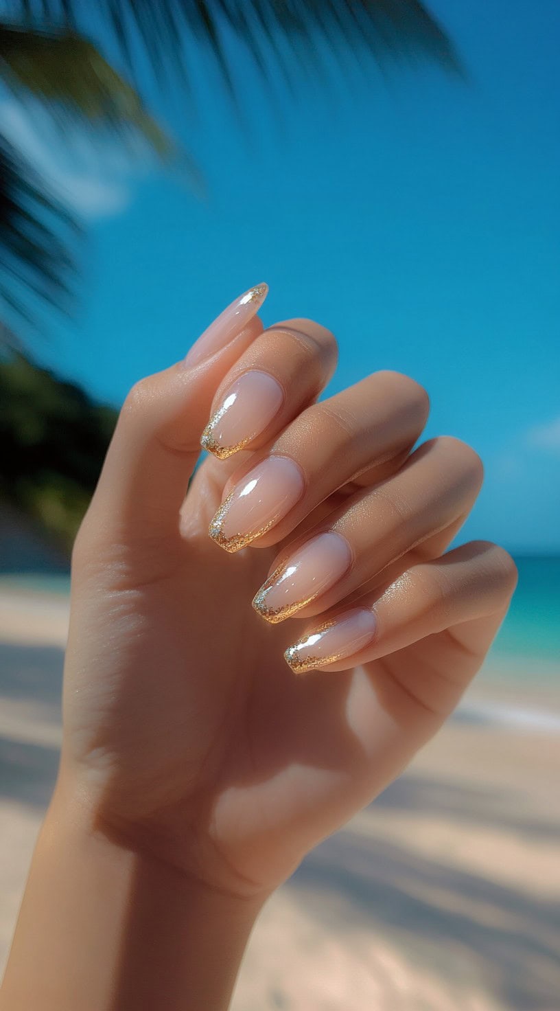 Almond-shaped nails with a glossy nude base and gold glitter tips, glistening in the sunlight.