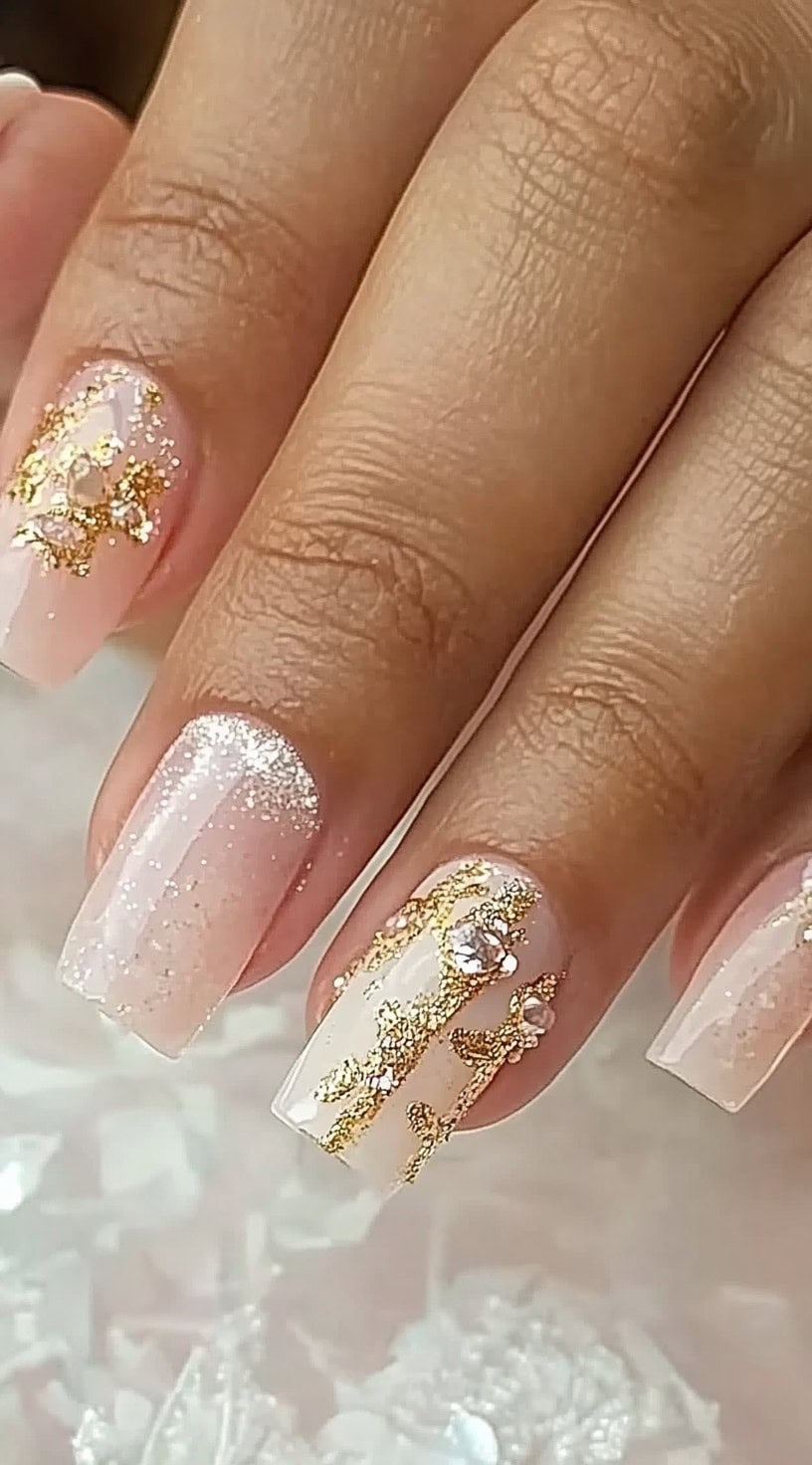 A set of coffin-shaped nails with a sheer nude base, gold lace detailing, and sparkling rhinestone embellishments.