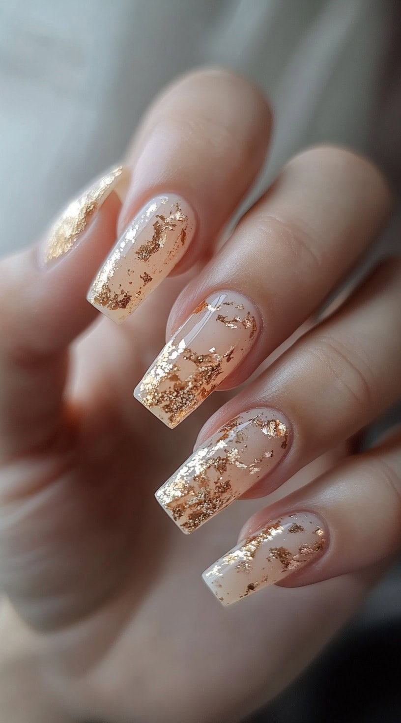 A hand with long coffin-shaped nails adorned with transparent nude polish and scattered gold leaf foil accents.
