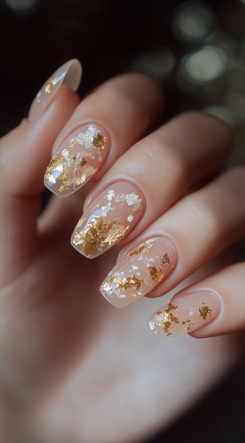 A close-up of medium-length, square-shaped nails with a nude sheer base, featuring luxurious gold leaf flakes scattered throughout.