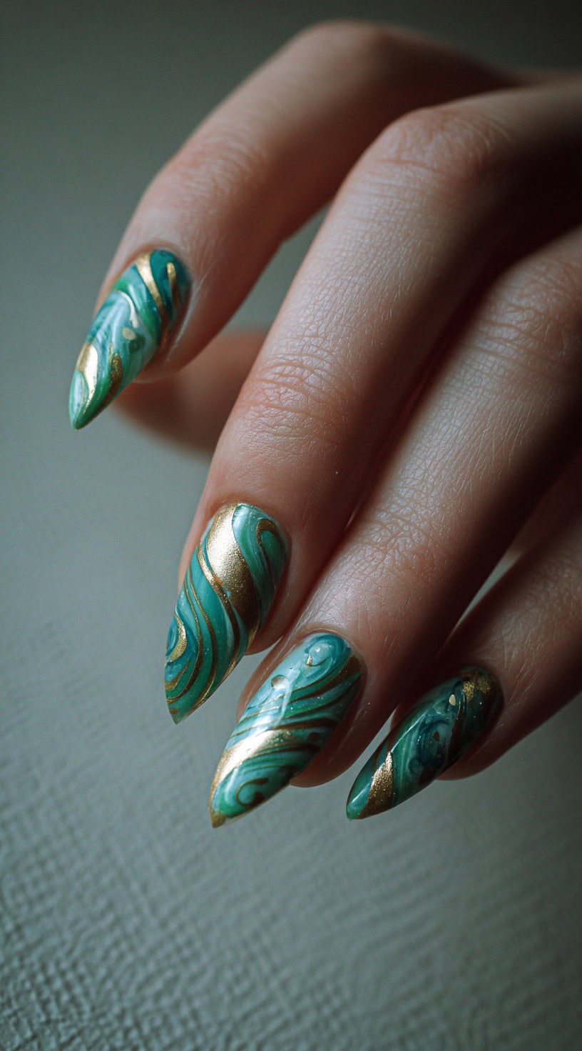 A hand with stiletto-shaped nails featuring deep jade marbling interwoven with sweeping gold metallic swirls.