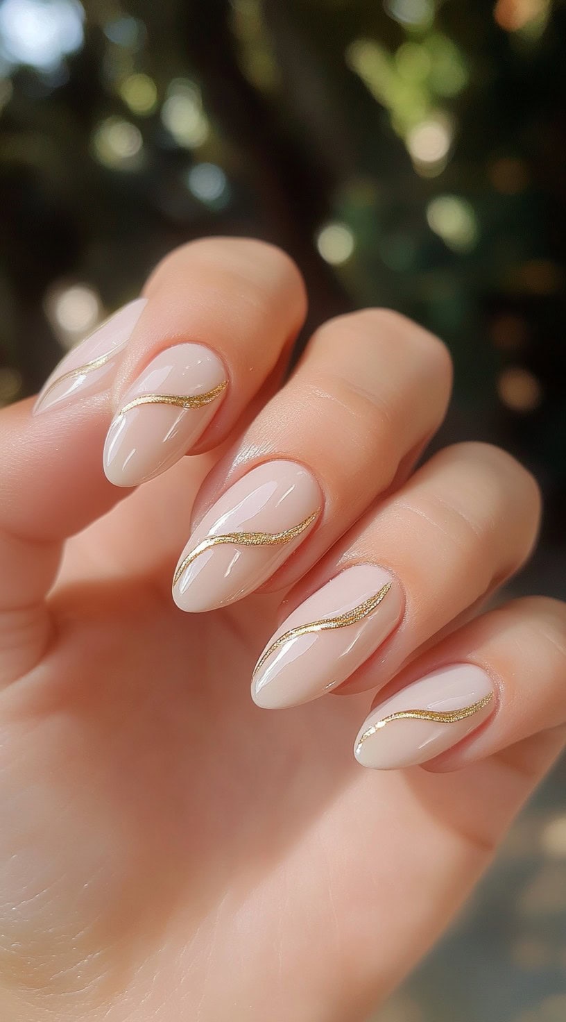 Hand with almond-shaped nude nails featuring delicate gold wave lines.