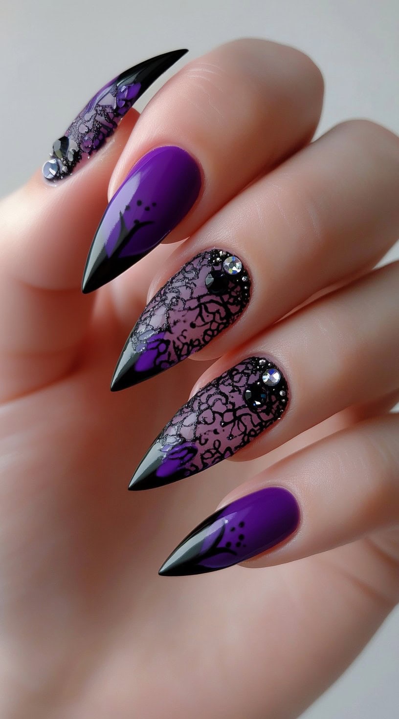 A hand with stiletto nails in purple and black featuring lace-like detailing, tiny rhinestones, and floral accents at the tips.