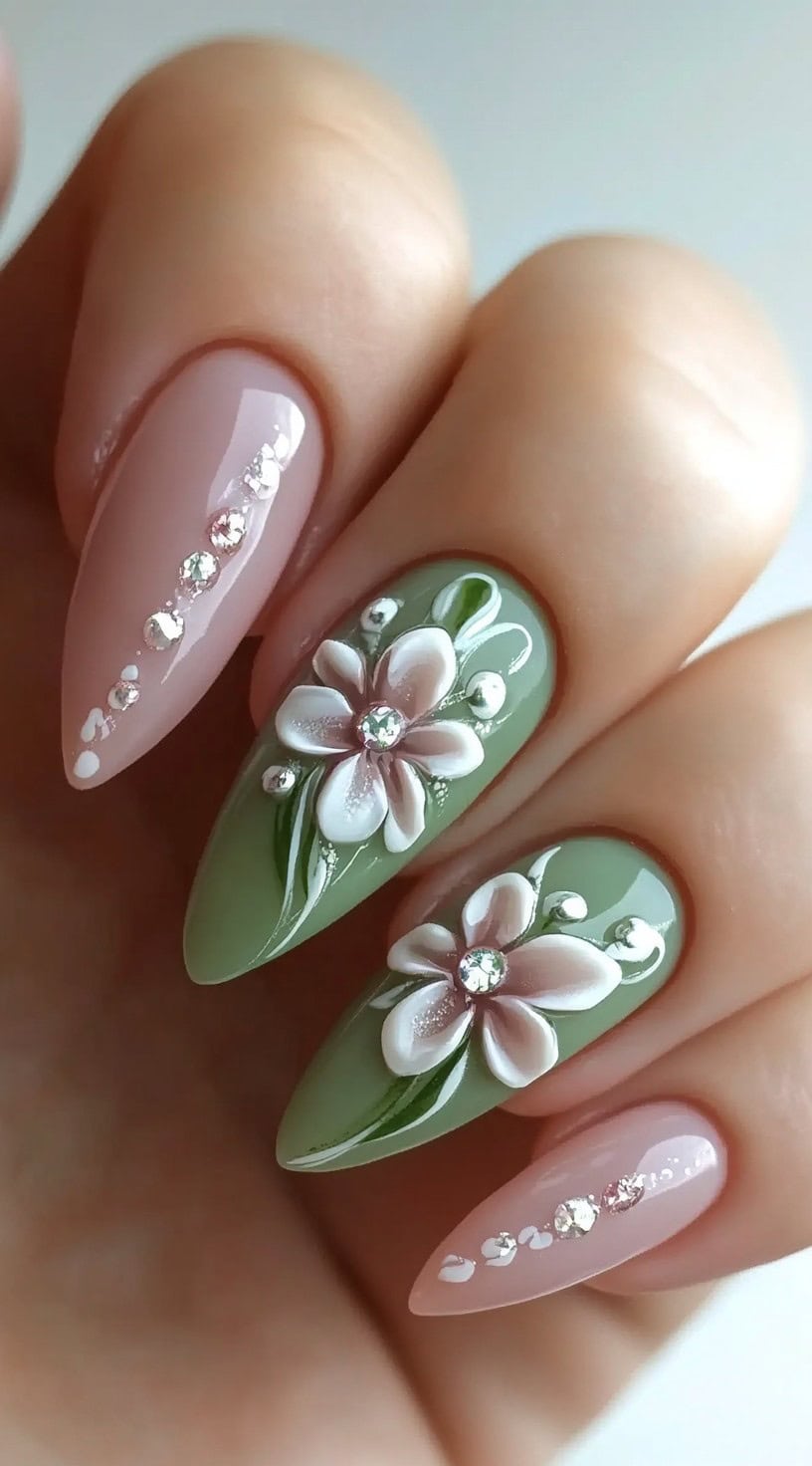 A set of almond-shaped nails featuring a soft sage green and nude base, decorated with white and pink 3D flowers, rhinestones, and delicate swirl accents.