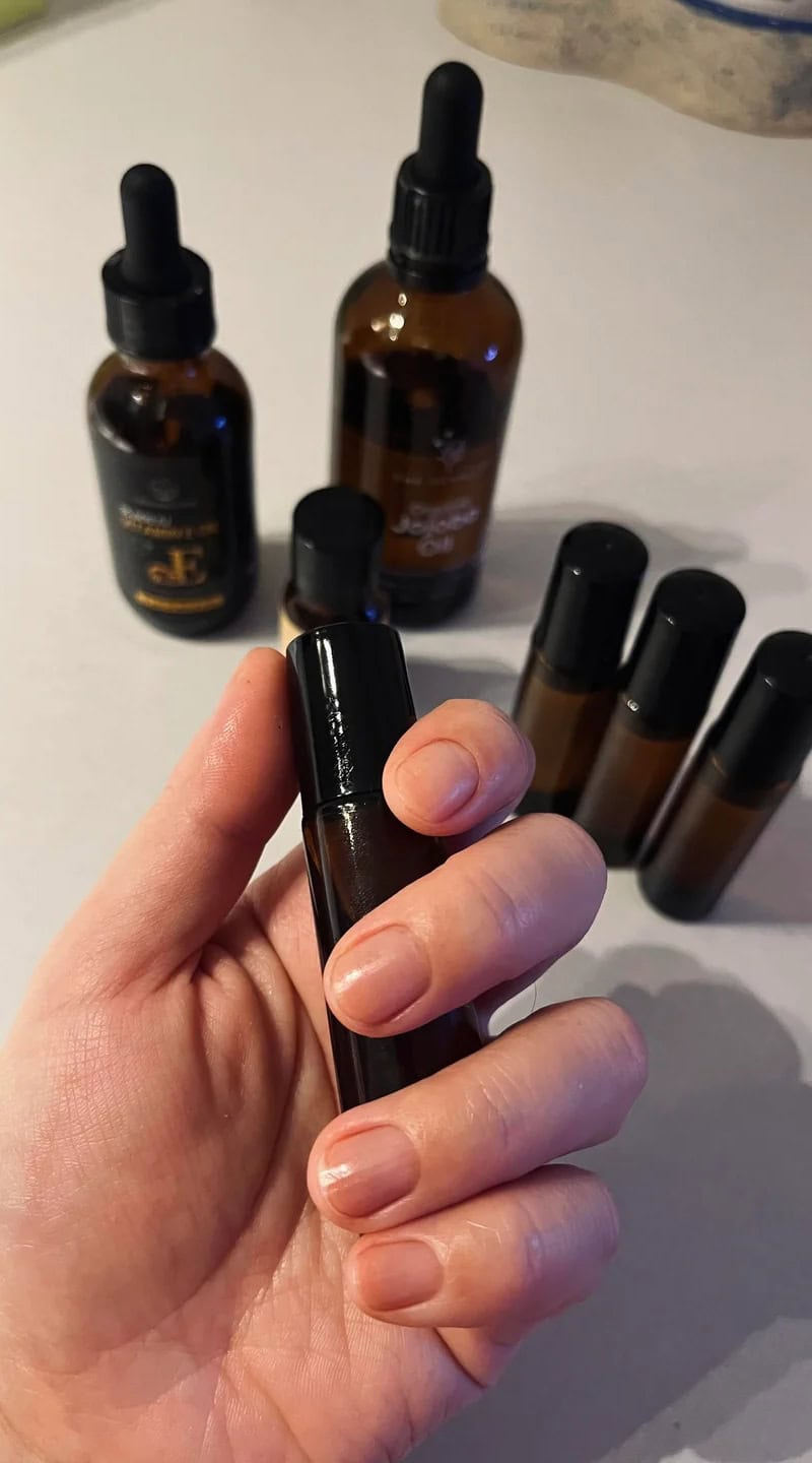 A hand holding a small amber glass bottle filled with homemade cuticle oil, with other DIY ingredients visible in the background.