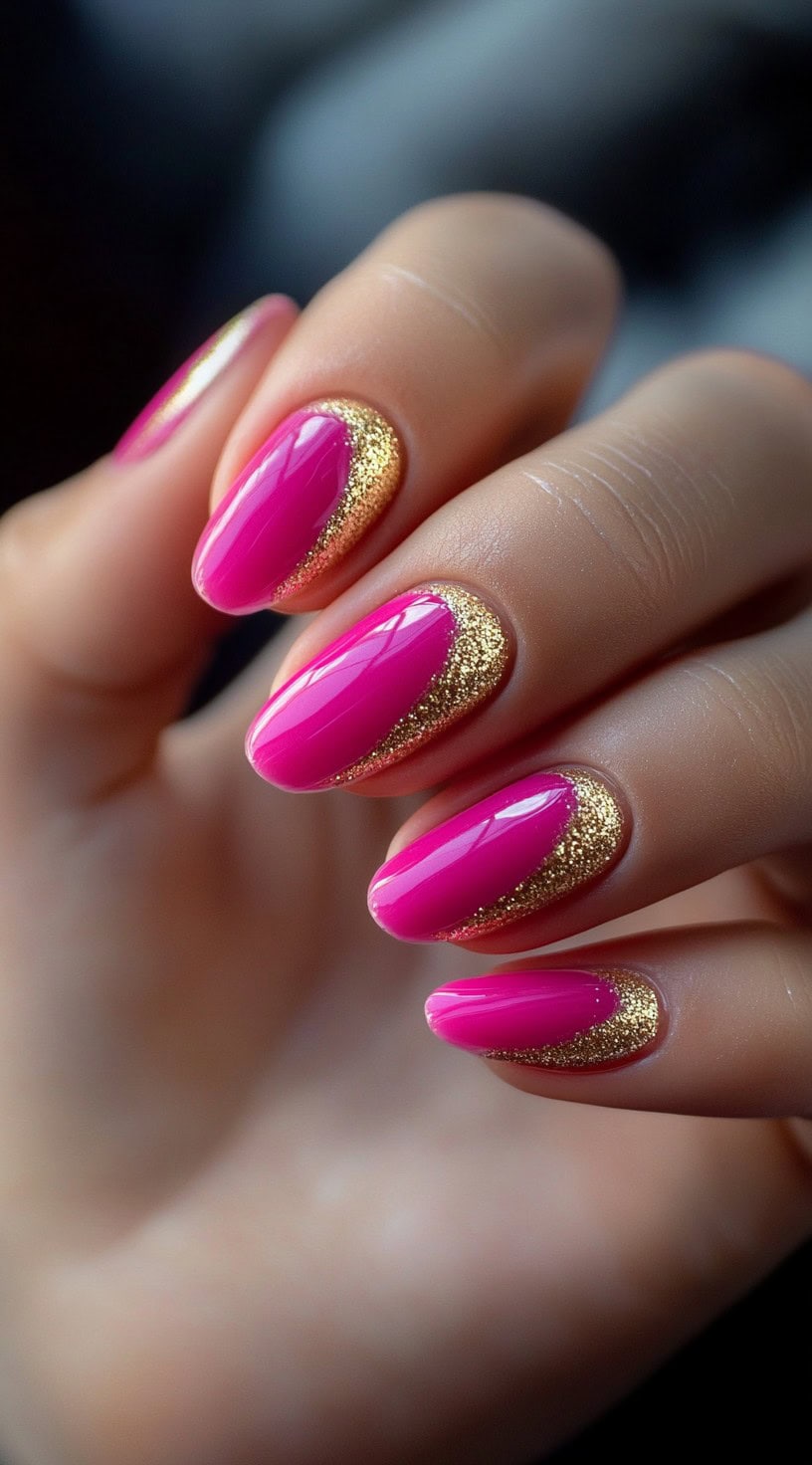 A set of almond-shaped nails painted in bold hot pink, featuring a crescent of gold glitter at the cuticle.