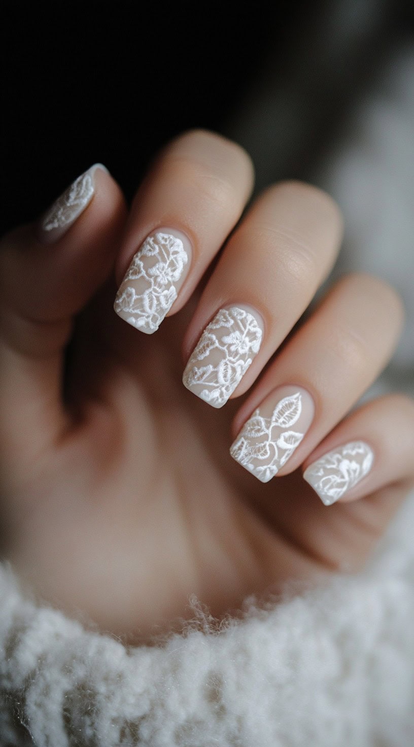 A close-up of a bride's hand with short square nails, coated in soft white and detailed with intricate lace-inspired designs for a romantic, bridal-inspired look.