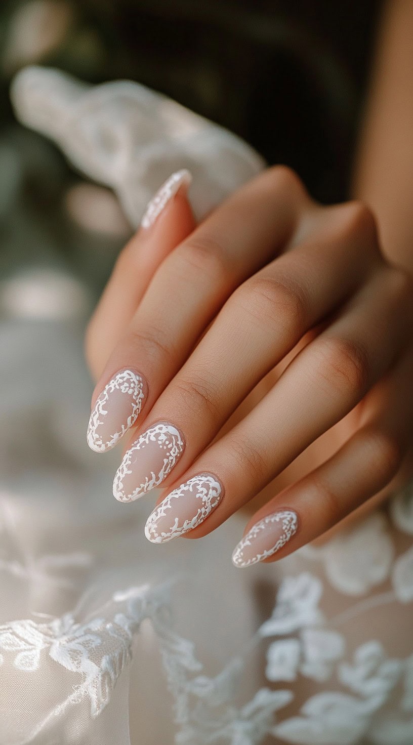 A hand with almond-shaped nails featuring intricate white lace-inspired designs over a sheer nude base.