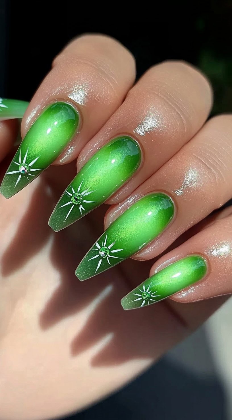 A close-up of long coffin-shaped nails with a lime green ombre effect, decorated with white starburst designs and small rhinestones at the center.