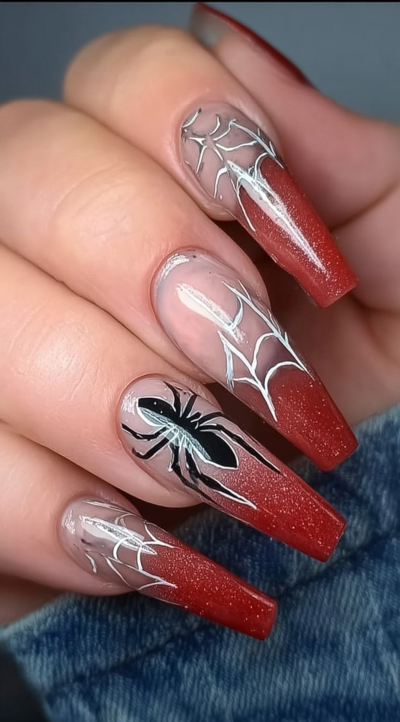 A hand with red ombre coffin nails featuring hand-painted white spiderwebs and a black spider on the ring finger.