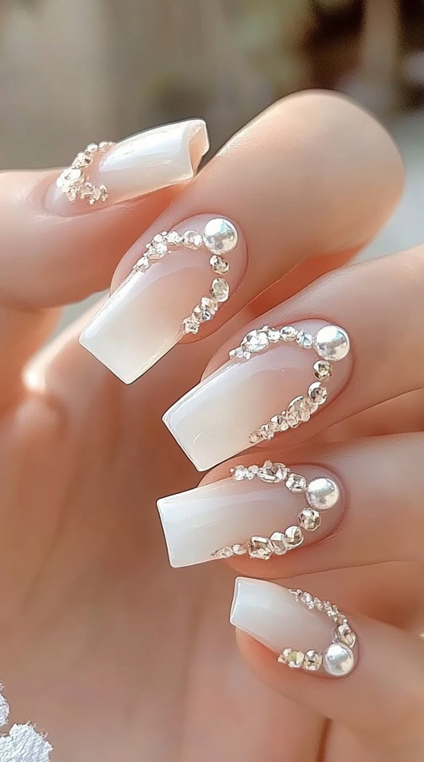 Square-shaped nails with a sheer white French tip, adorned with curved pearl and crystal embellishments.