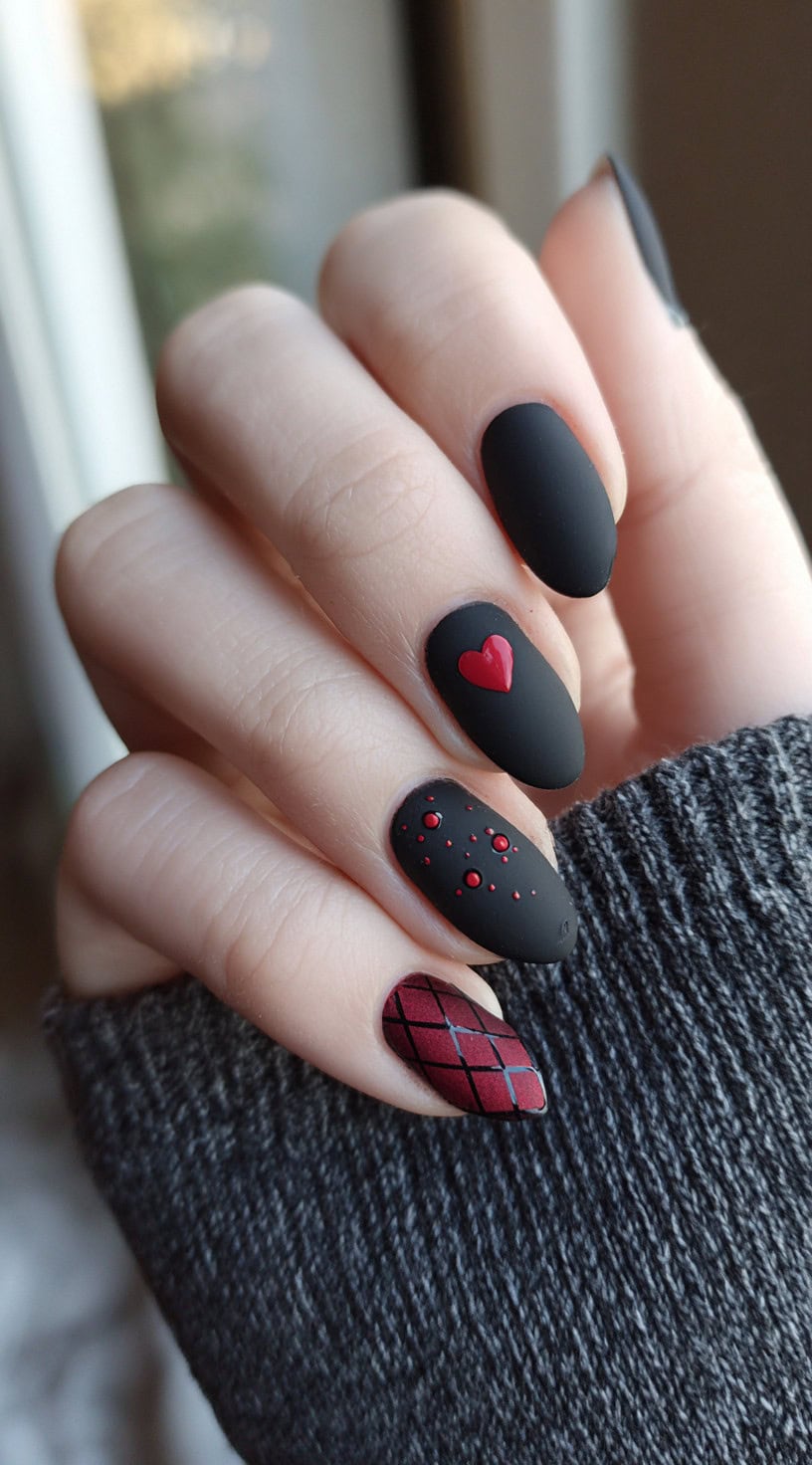 Almond-shaped matte nails featuring solid black, a single red heart accent, red dot clusters, and a burgundy harlequin diamond pattern.
