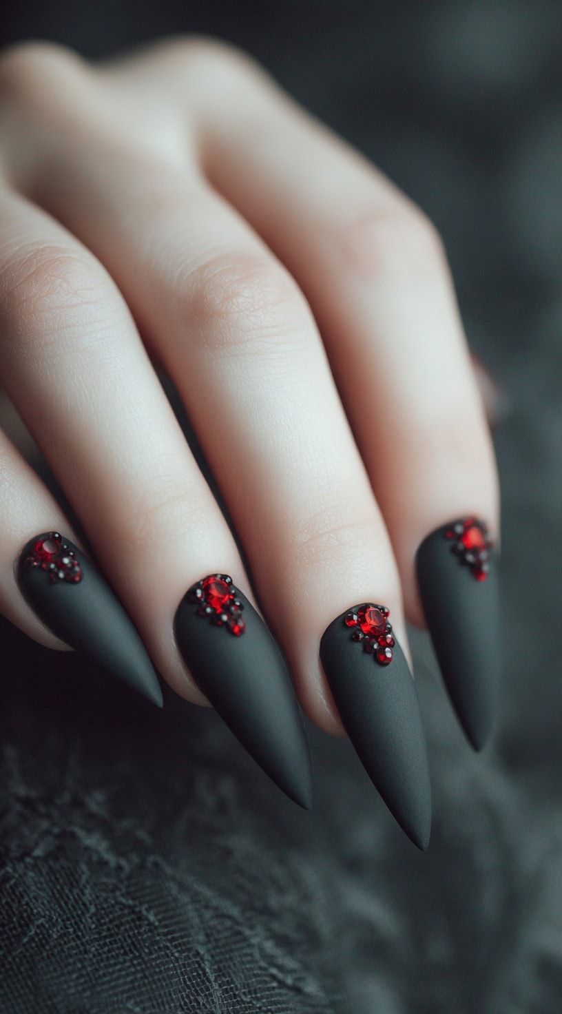 Matte black stiletto nails topped with small clusters of blood-red rhinestones at the cuticle.