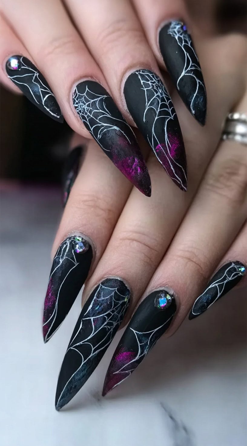 A hand with stiletto-shaped matte black nails featuring galaxy-style color splashes, spider webs, and rhinestone accents.