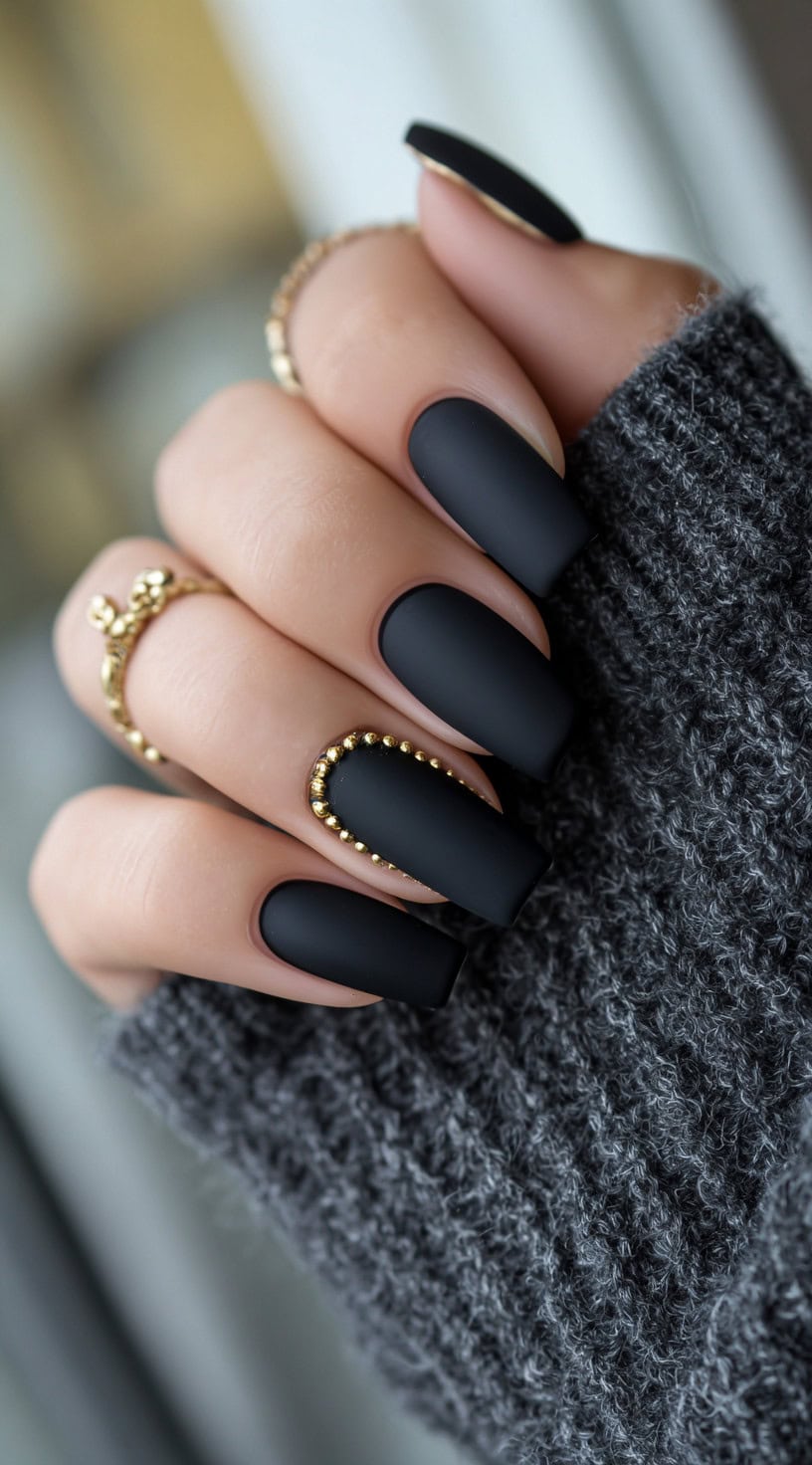 A hand with matte black square nails featuring gold bead embellishments outlining one accent nail.