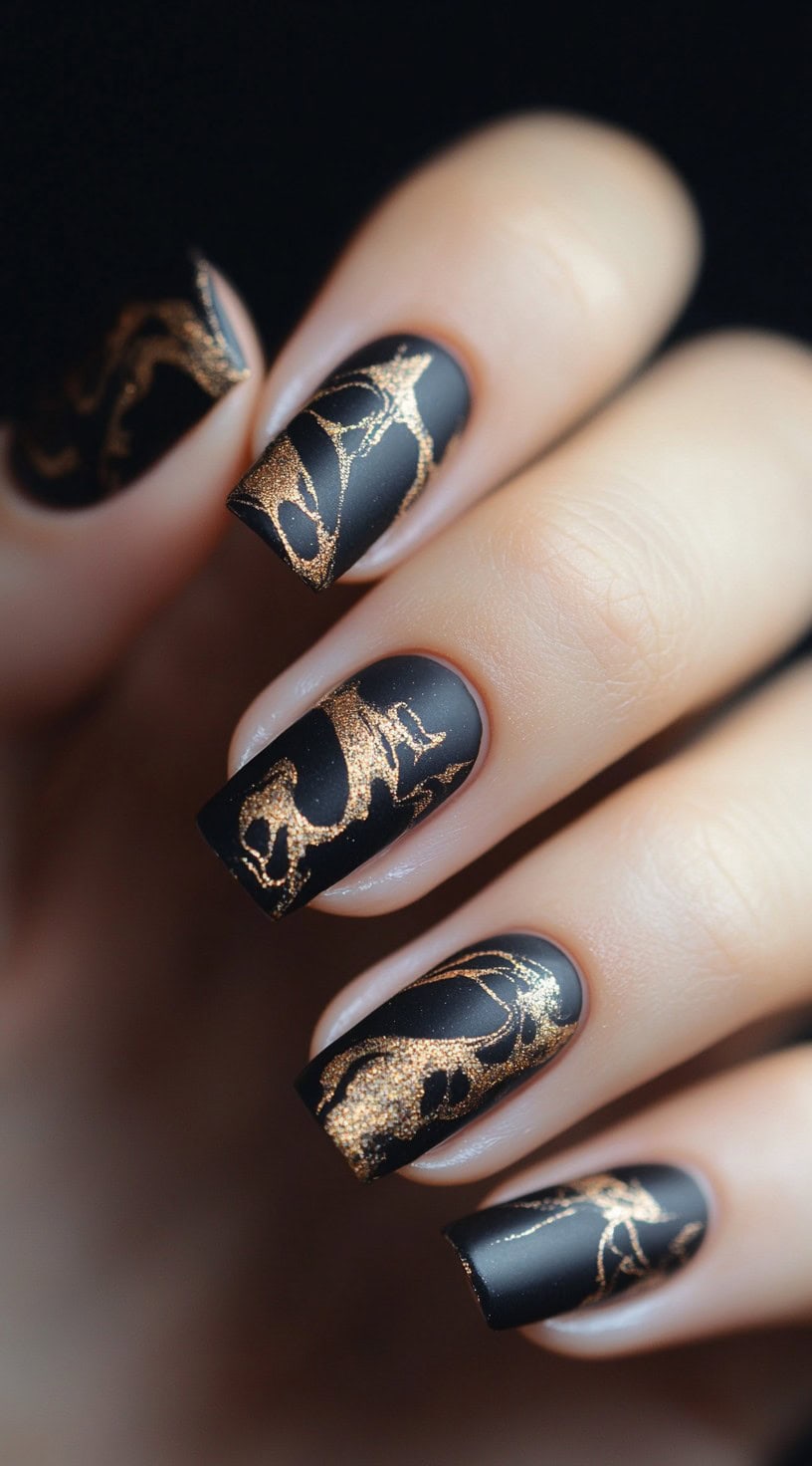 A hand with square-shaped nails painted in matte black, featuring gold foil detailing in an abstract, artistic pattern.
