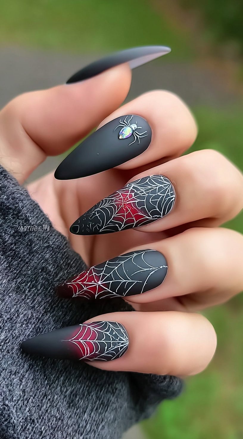 A hand with stiletto-shaped nails featuring a matte black base, red gradient tips, white spiderwebs, and a gem-like spider accent.