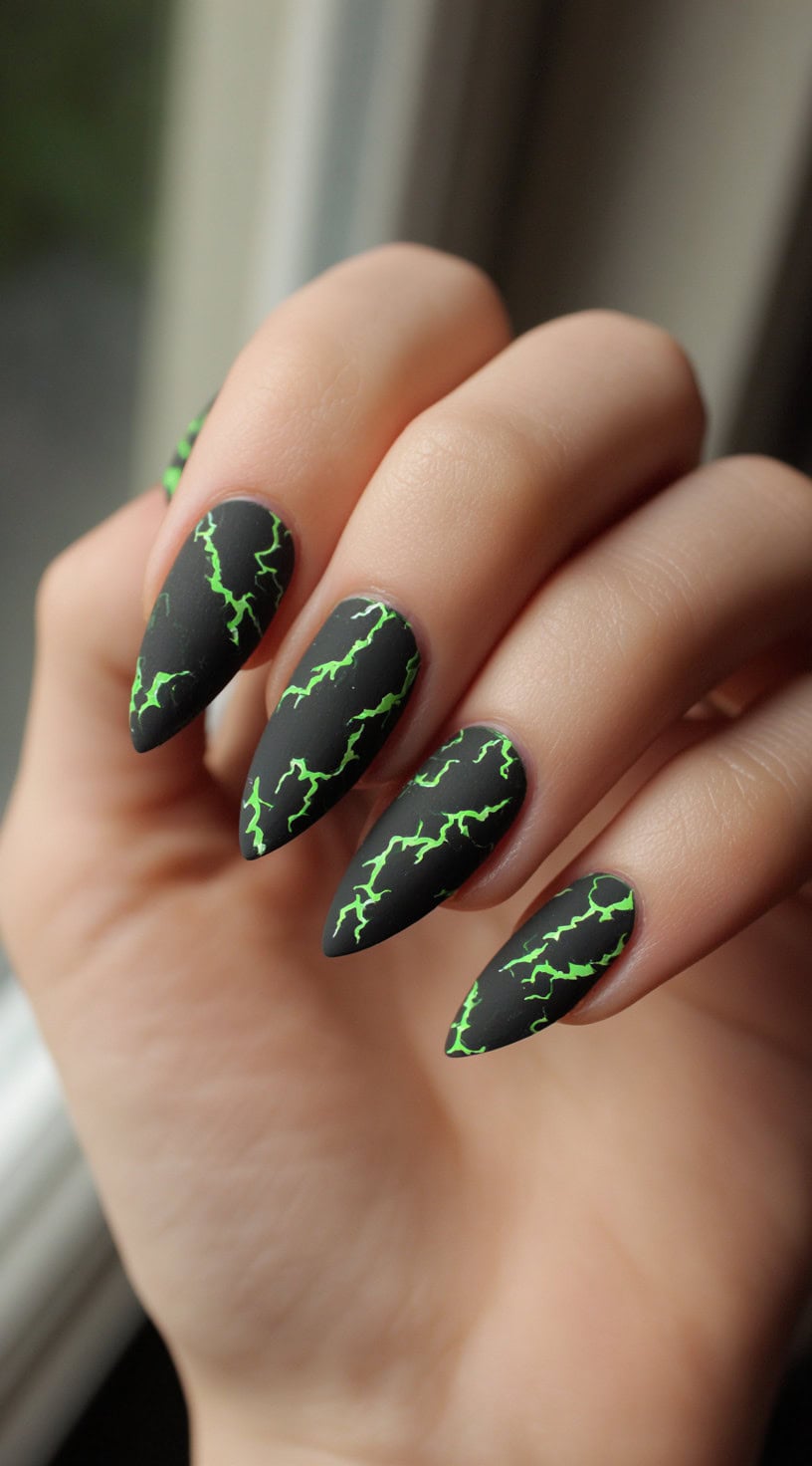 Stiletto nails painted matte black with neon green lightning bolt crackle patterns running across each nail.