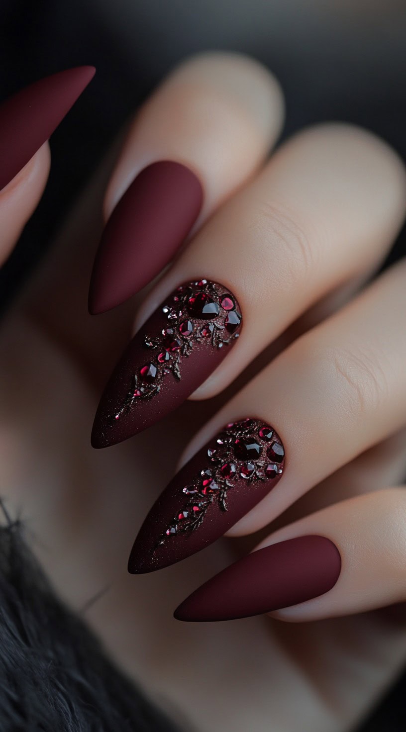 Matte burgundy stiletto nails adorned with deep red gem clusters forming vine-like patterns.