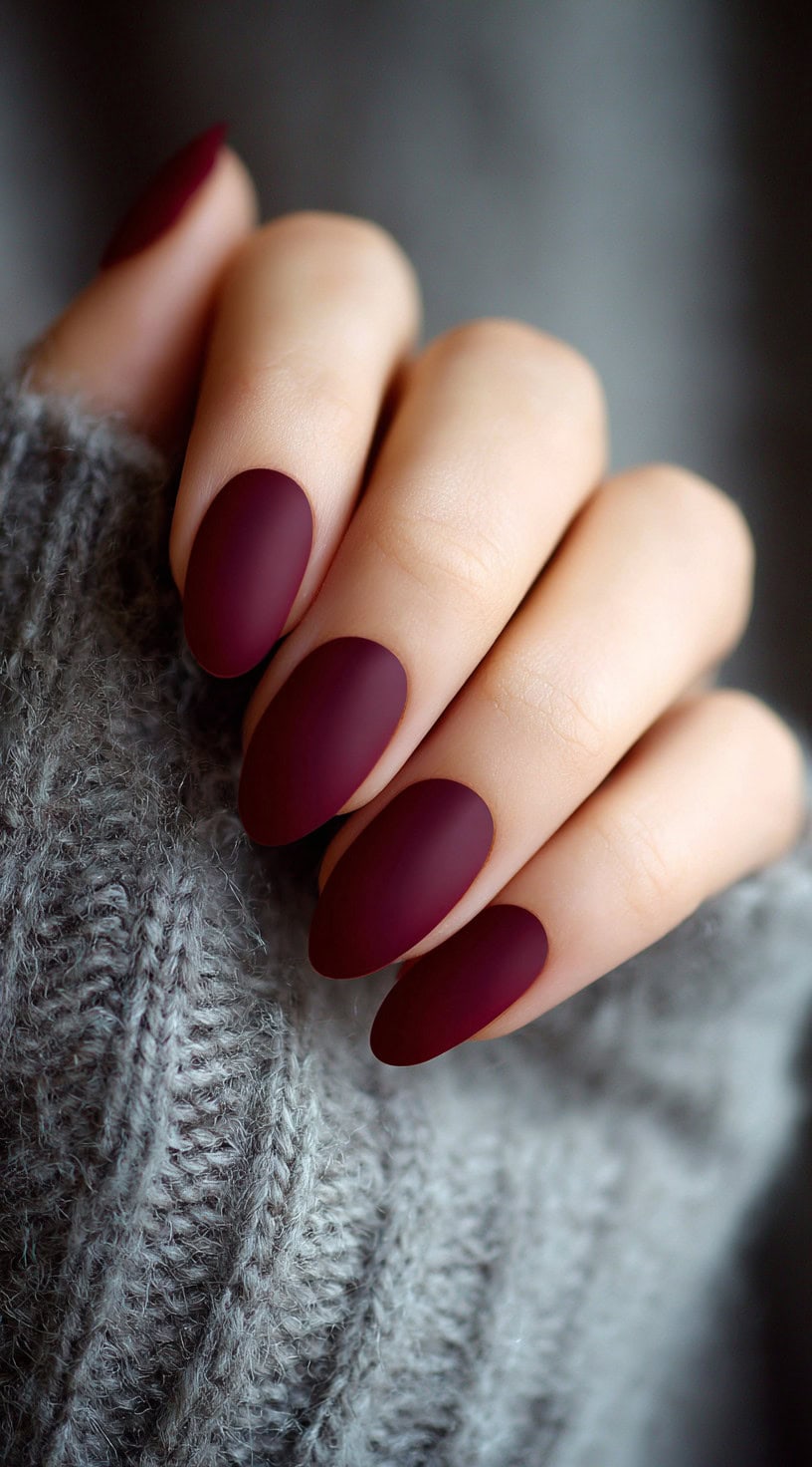 A hand with oval-shaped matte burgundy nails peeking through a soft gray sweater.
