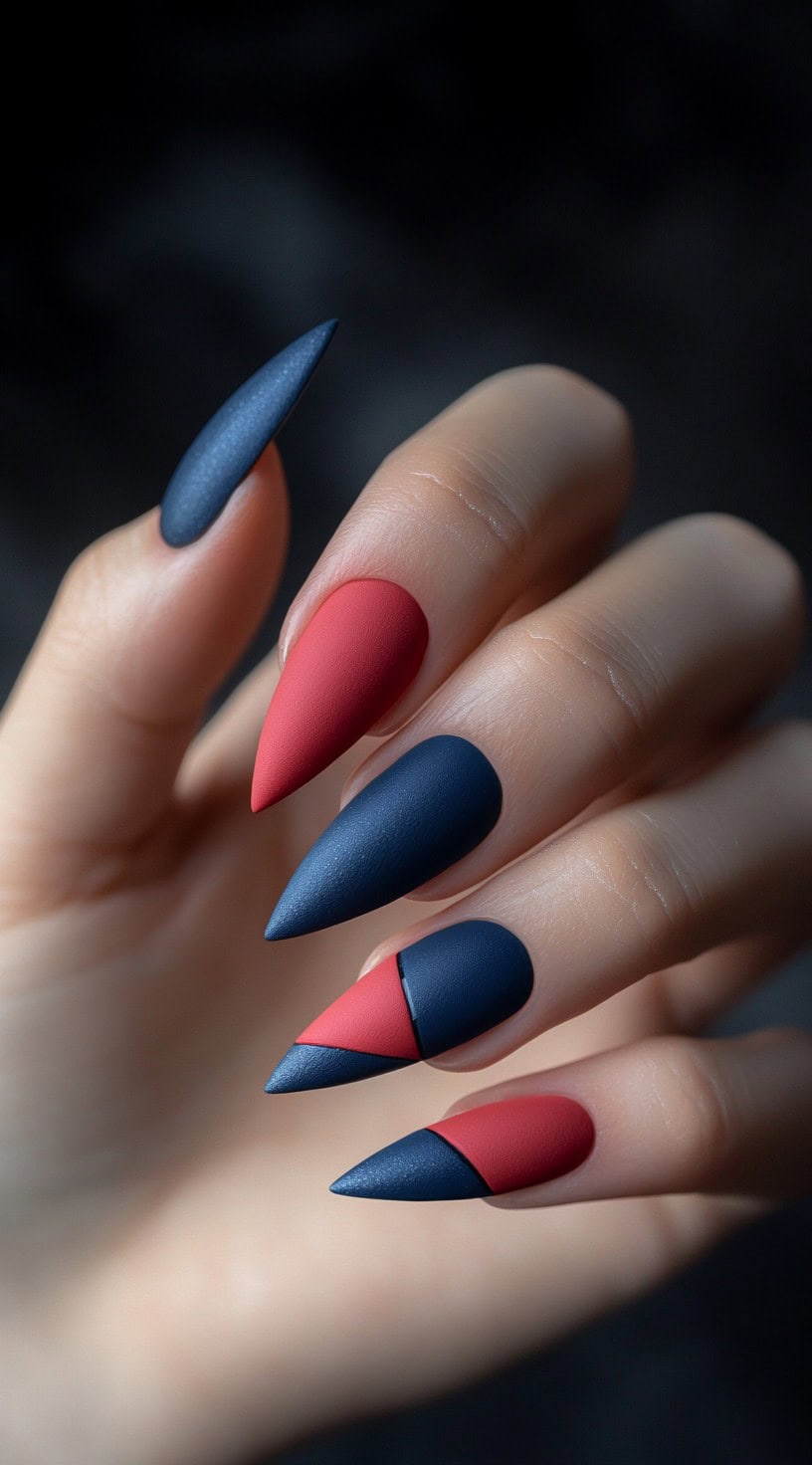 A hand with long, stiletto-shaped nails featuring a bold matte red and blue geometric pattern, with each nail divided into angular sections.