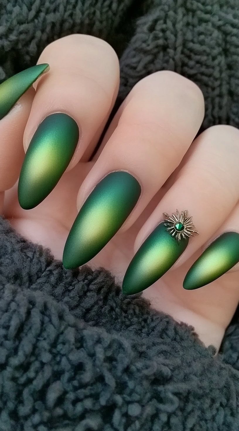 A set of long almond-shaped nails featuring a deep green matte ombre effect, with a gold sunburst charm on the accent nail.