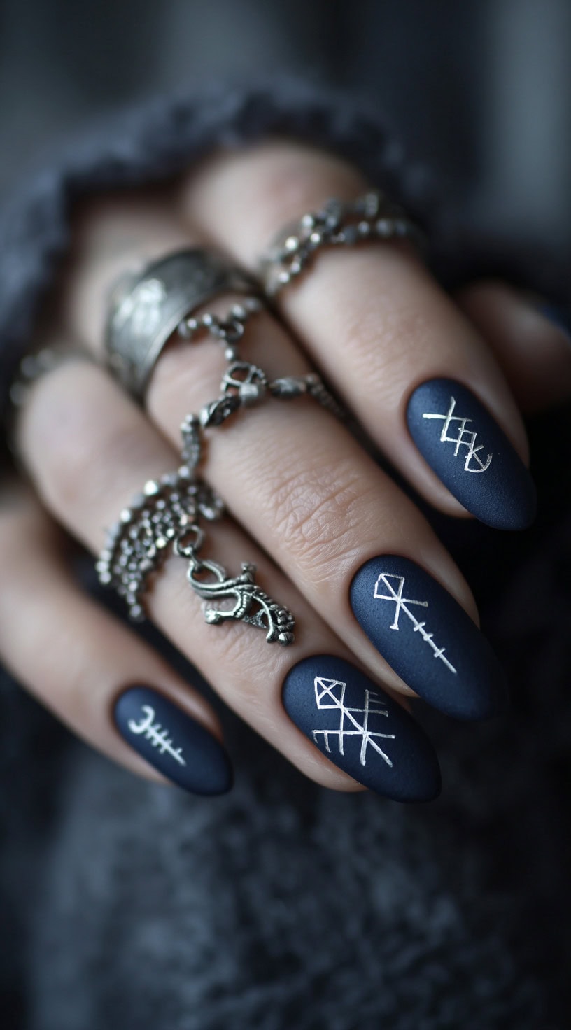 A hand with almond-shaped matte navy blue nails featuring clean white rune symbols and silver Viking rings.