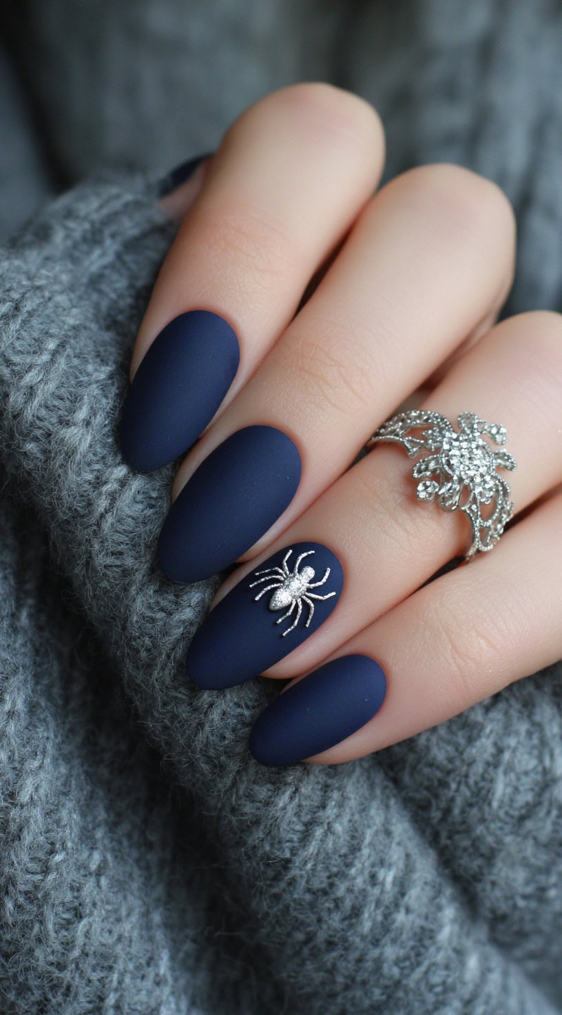 Almond-shaped matte navy nails with a textured silver spider embellishment on the ring finger.