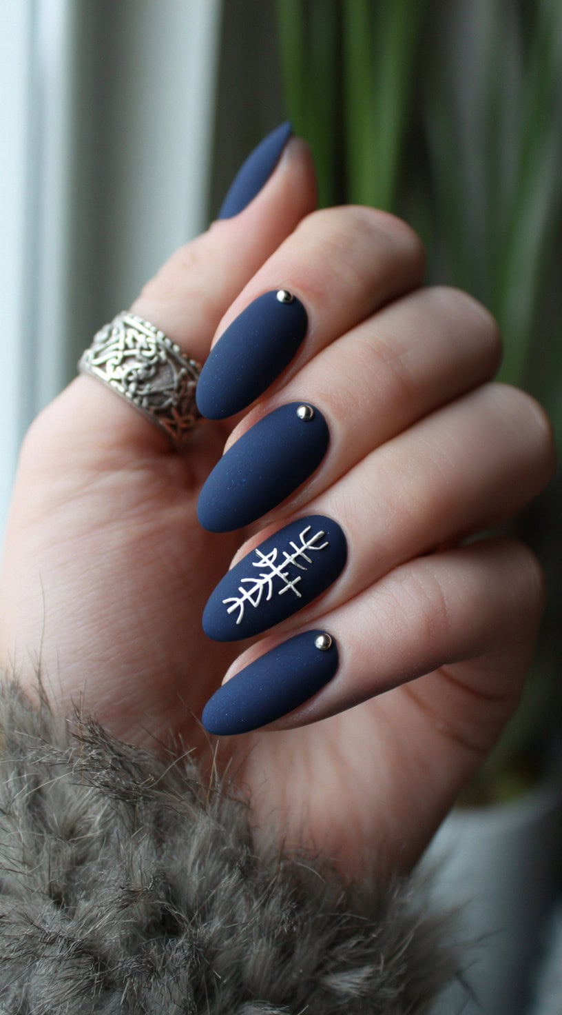 A hand with long almond matte navy nails featuring silver rune designs and tiny metallic studs at the base.