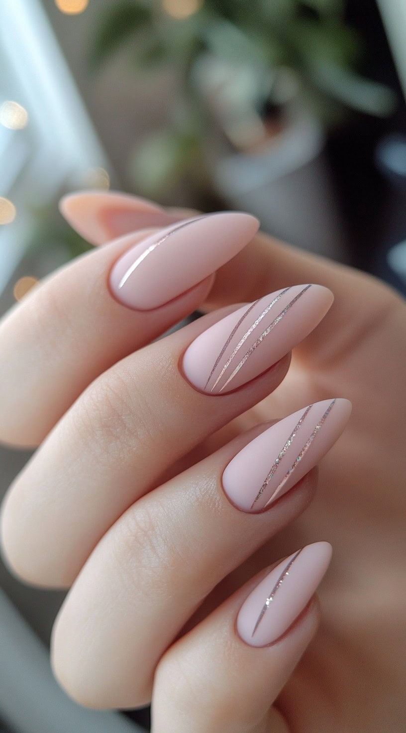 A hand with almond-shaped nails in a soft matte pink shade, featuring fine silver metallic stripes running diagonally across select nails.