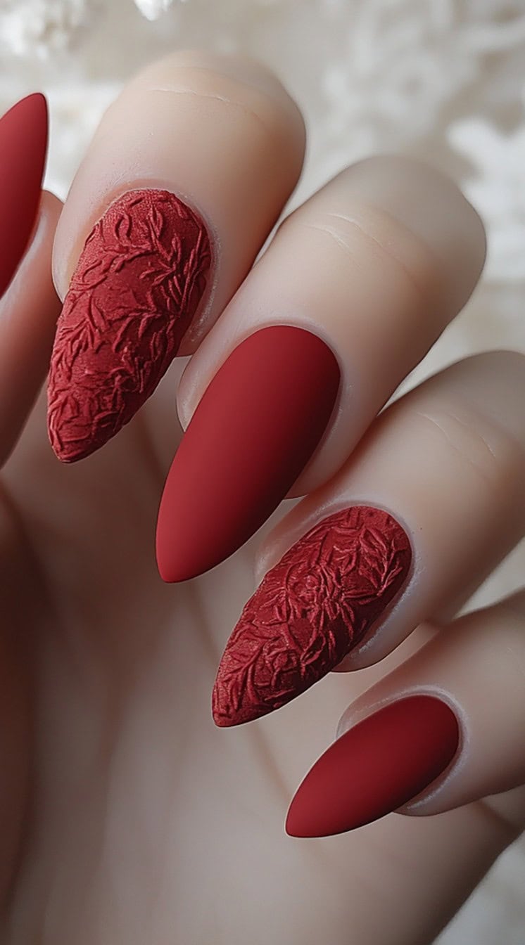 A set of matte red stiletto nails featuring a soft velvet texture on select nails, with embossed floral patterns for an elegant and tactile finish.