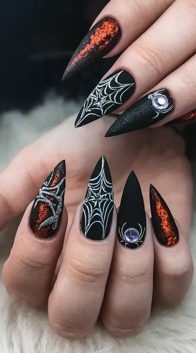A hand with matte black stiletto nails featuring glowing lava accents, spider webs, and small gem embellishments.