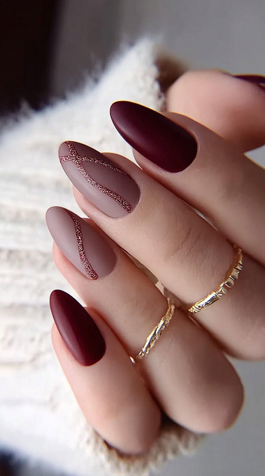 A hand with matte almond nails alternating between wine and taupe, with the taupe nails featuring fine glitter lines in deep red.