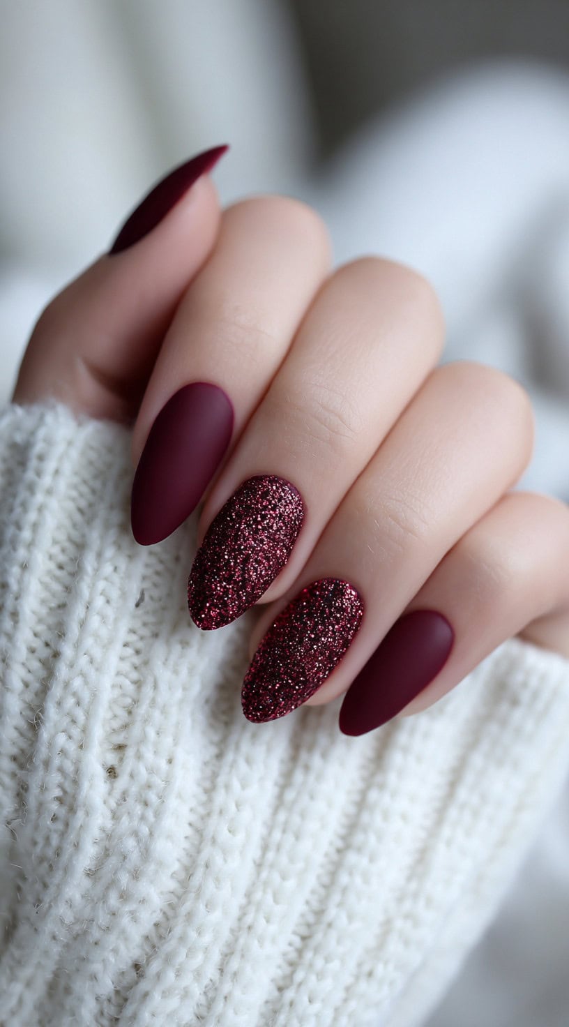 A hand with matte burgundy almond nails and two accent nails fully covered in wine red glitter.