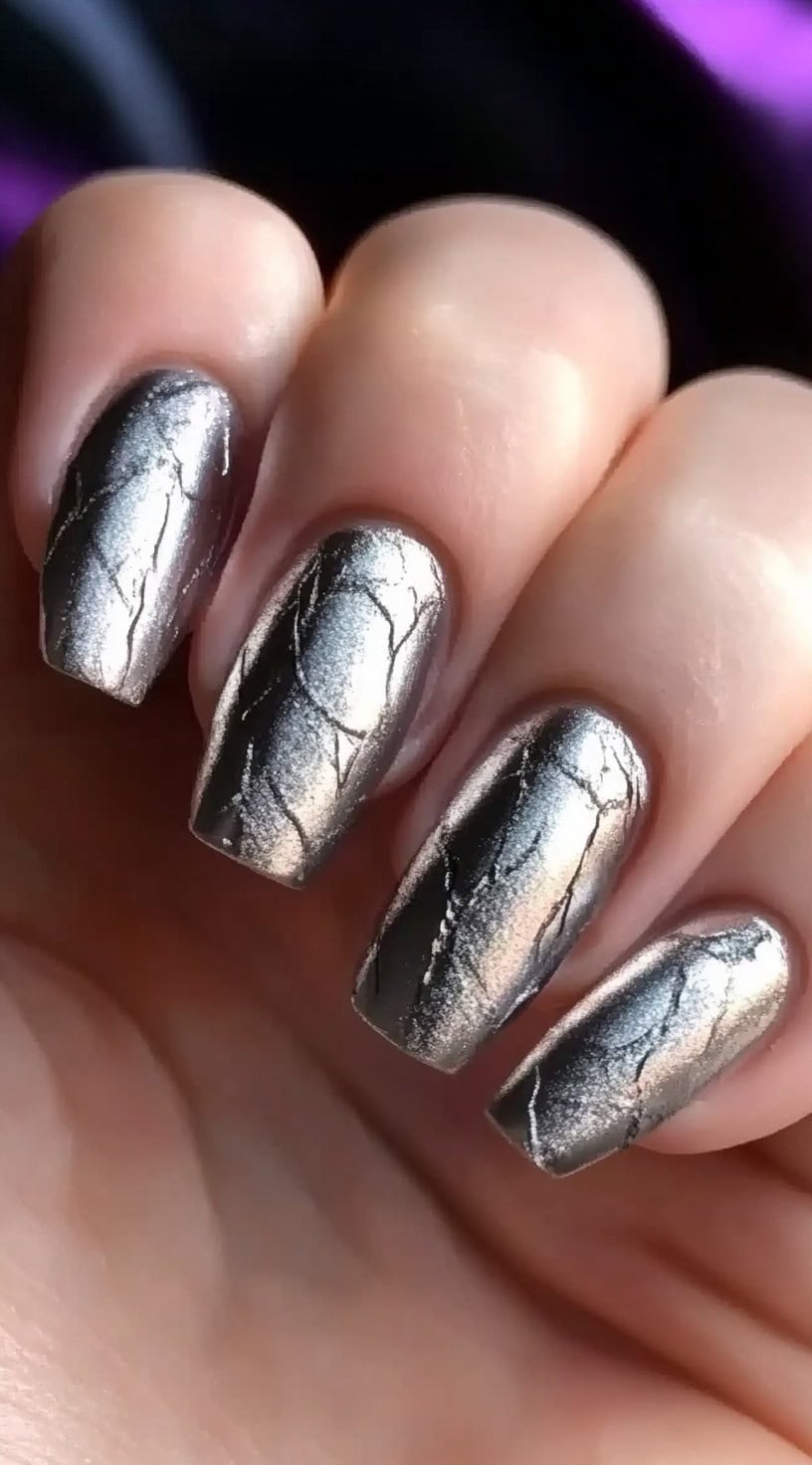 A close-up of nails painted in a metallic silver shade with an eye-catching cracked effect, resembling shattered chrome.