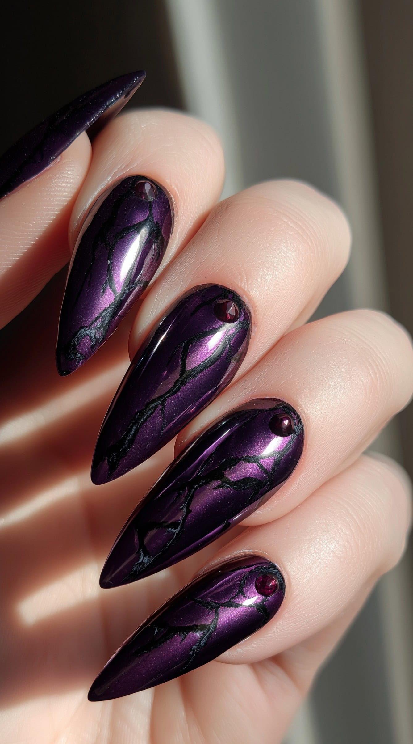 A hand with long metallic purple stiletto nails, black cracked-vein designs, and small blood-red gemstone accents near the cuticles.