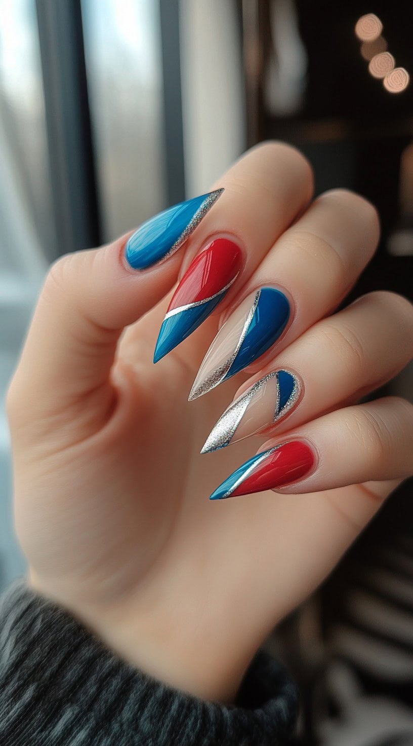A hand with long, sharp stiletto nails featuring a metallic red and blue design accented with silver diagonal stripes.