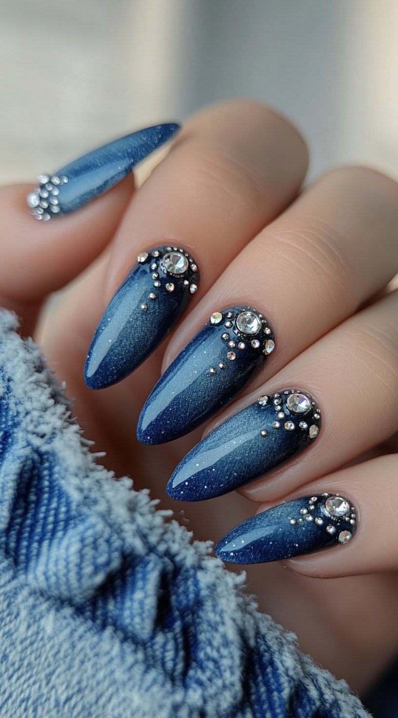 Long, almond-shaped nails painted in deep midnight blue with a subtle shimmer effect, accented by crystal embellishments along the cuticles.