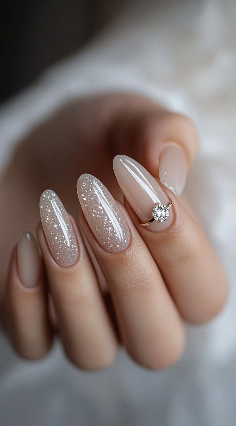 Five oval nails with a milky nude base, soft glitter on two nails, and a delicate ring-inspired charm on the middle finger.
