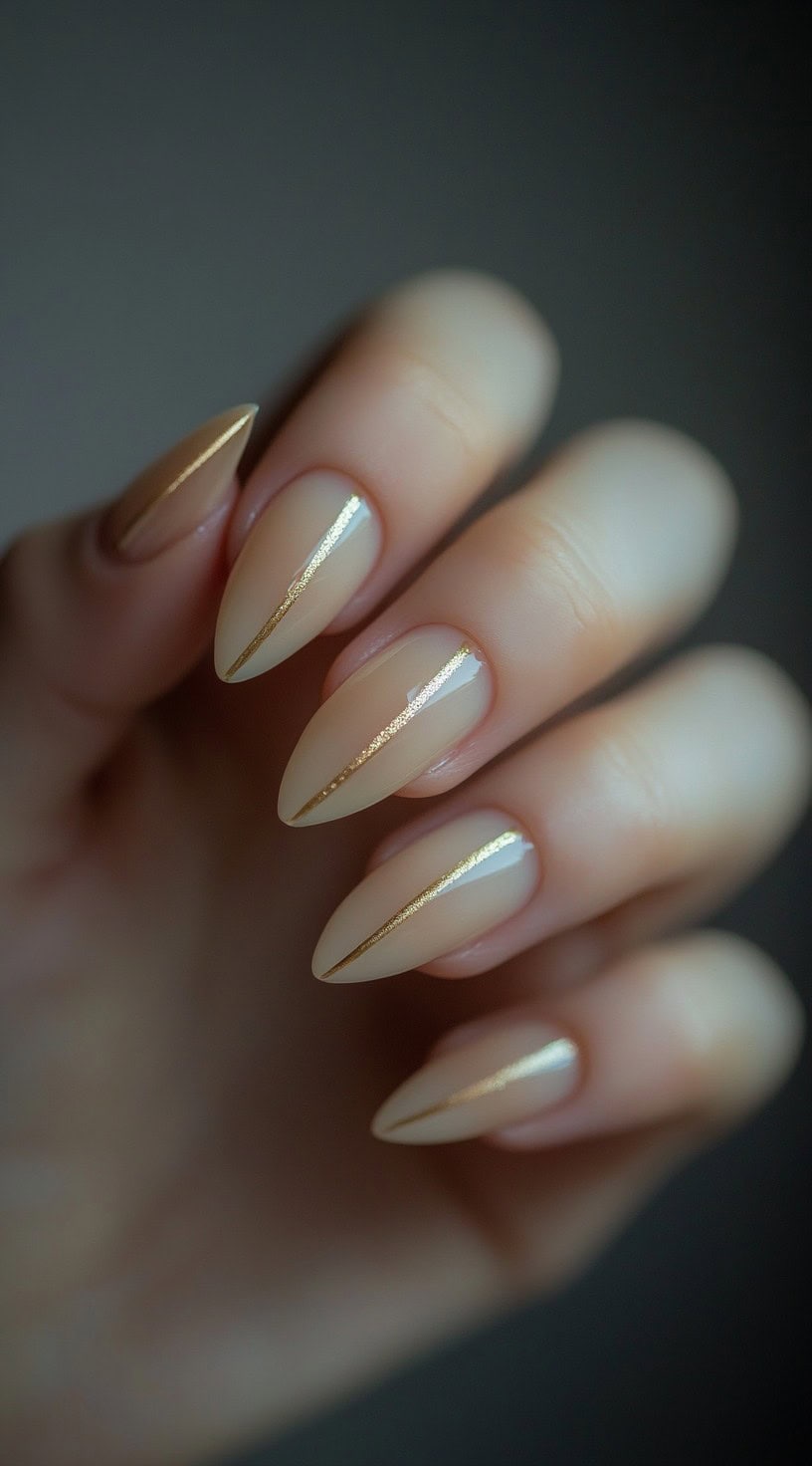 A hand with almond-shaped nails painted in a soft nude shade, featuring thin, precise gold stripes running vertically down each nail.