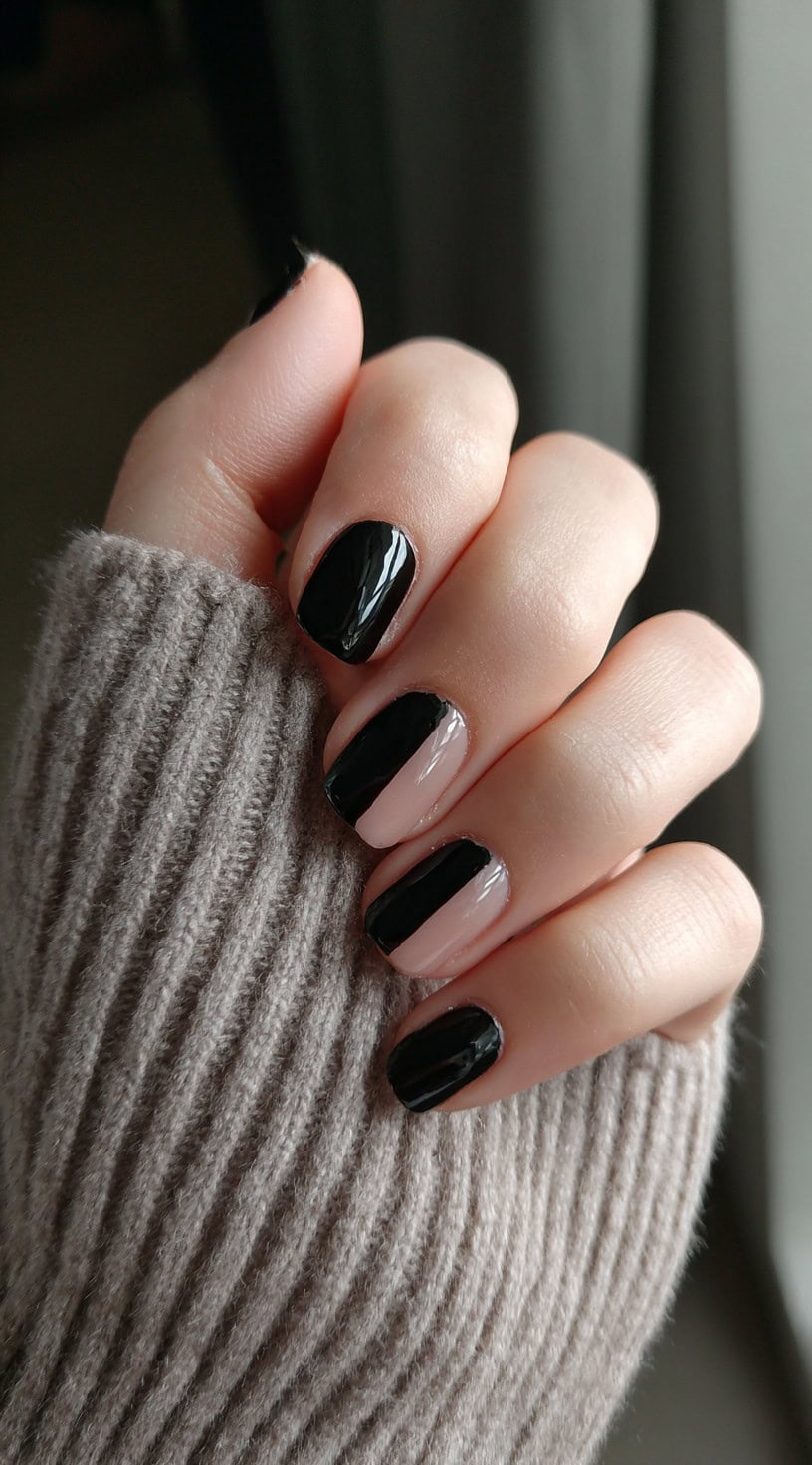 A hand with short nails painted with a vertical half-and-half design using black and clear nude polish.