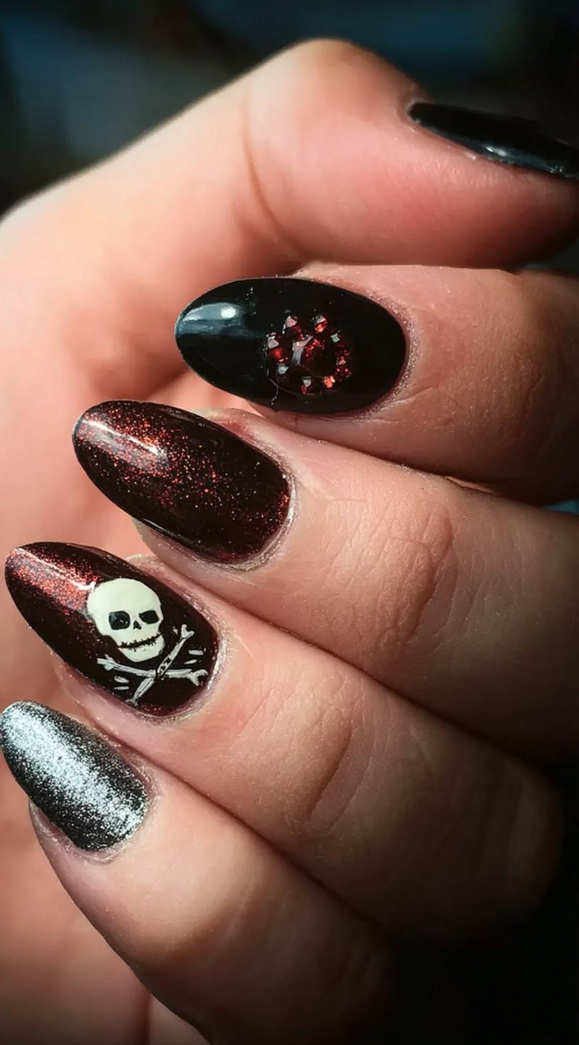 Almond nails alternate between black, red shimmer, silver chrome, and skull accents with gemstone detailing.