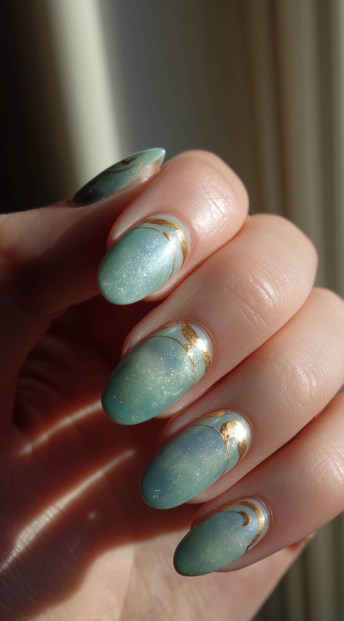 A hand with almond-shaped nails in a soft, glittery jade hue accented by gold crescent moon shapes near the cuticles.