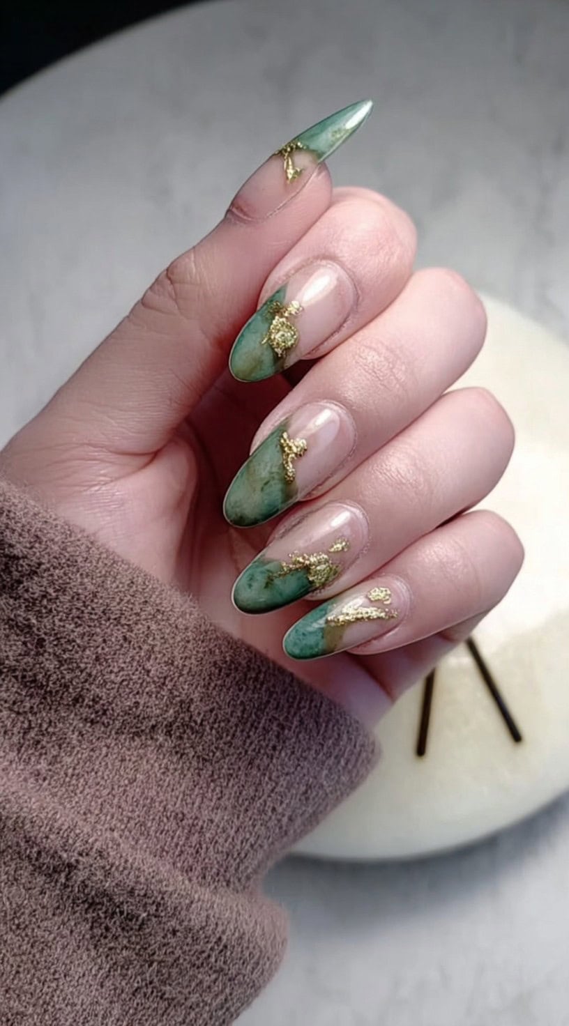 A hand with almond-shaped nails featuring sheer nude bases and jade marble tips accented with irregular gold foil patches.