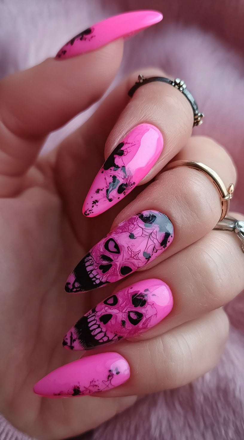 Hand with neon pink almond nails showcasing black skull designs and splatter patterns.