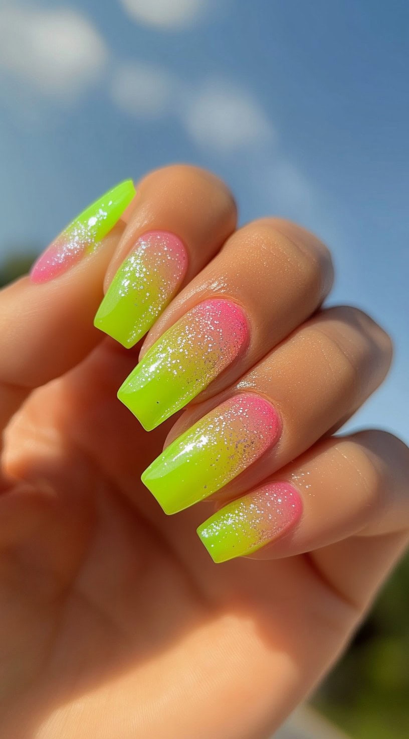 A hand with square-shaped nails featuring a hot pink and neon green glitter ombre design under sunlight.
